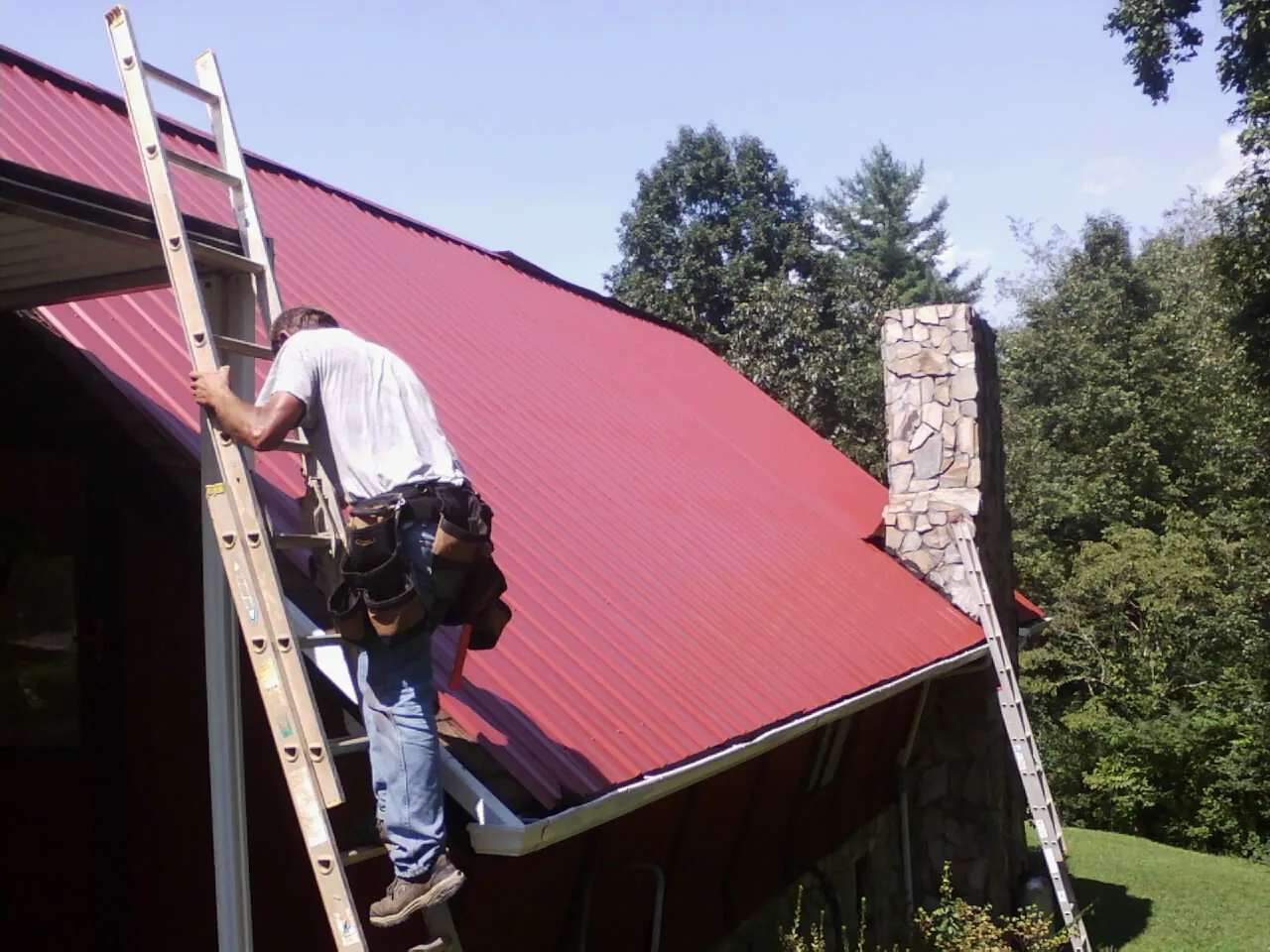 Roofer on a ladder beside a red metal roof and stone chimney — Soffit & Fascia Repair by Roofing Brothers in Green Lake
